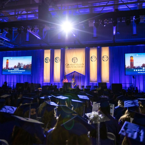 A stage with Grand Valley banners hanging over a blue curtain. A large crowd of graduates in graduation caps and gowns.