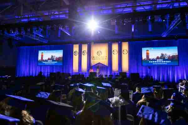 A stage with Grand Valley banners hanging over a blue curtain. A large crowd of graduates in graduation caps and gowns.