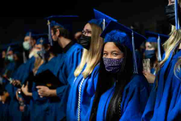 A graduate in a blue cap and gown wears a mask and poses for a photo.