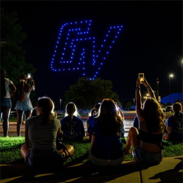 People sit on the ground at night to watch a drone show. The drone is spelling out a blue GV.