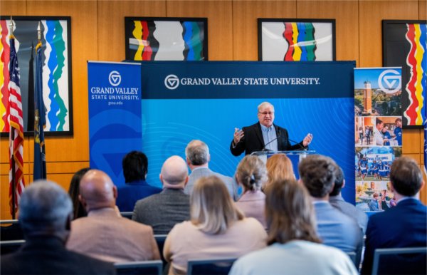 A person standing at a podium gestures with arms open. Banners with the words Grand Valley State University, and the university logo, are in the background.