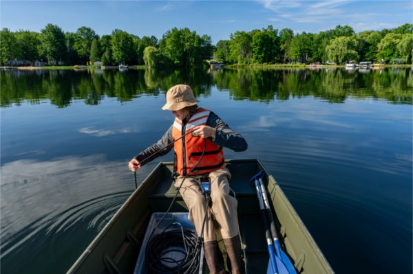 A person wearing a hat and life jacket dips a measuring device into the water while sitting in a small boat.
