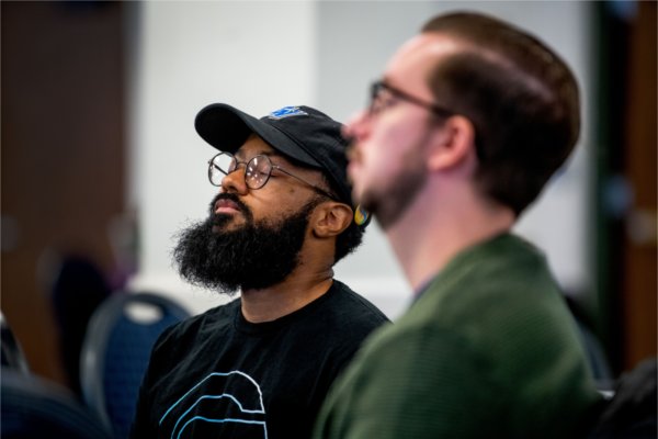 Two people closed their eyes as they meditate. One is wearing a hat with the letters, "GV."