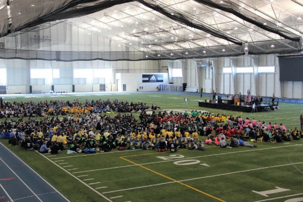 A large group of people sits on a floor with football-like yard markers. There also is a podium where people are sitting and one person is talking.