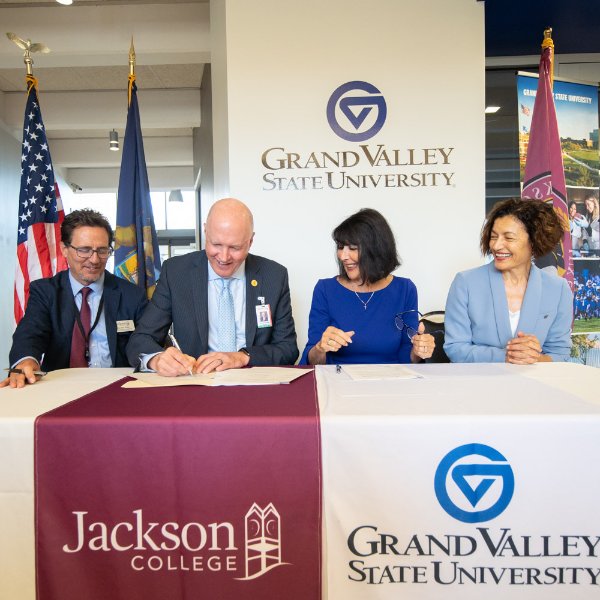 Four people smile while sitting at a table. The tablecloth has the words "Jackson College" and "Grand Valley State University."