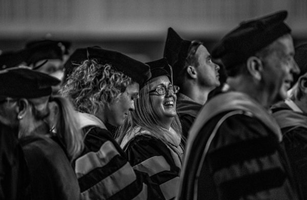  A person in academic regalia looks up and smiles amongst a sea of other people in regalia around them.