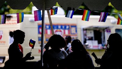 People stand in shadow below a banner of pride flags handing under a tent.