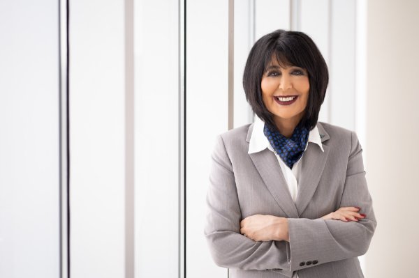 President Mantella standing with arms crossed in a hallway, wearing a gray suit and blue scarf