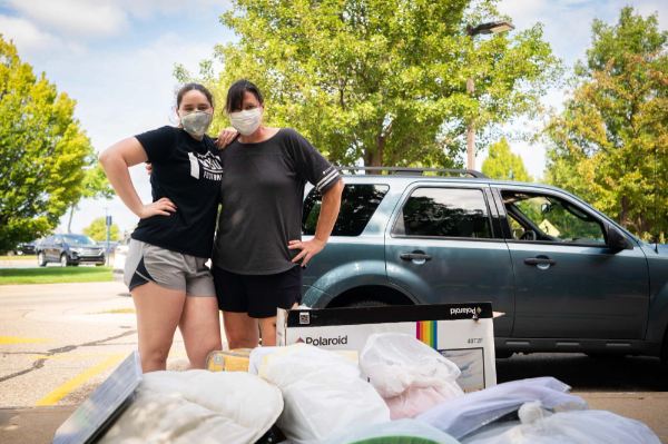 Two people stand with their arms around each other, posing behind a pile of someone's belongings.