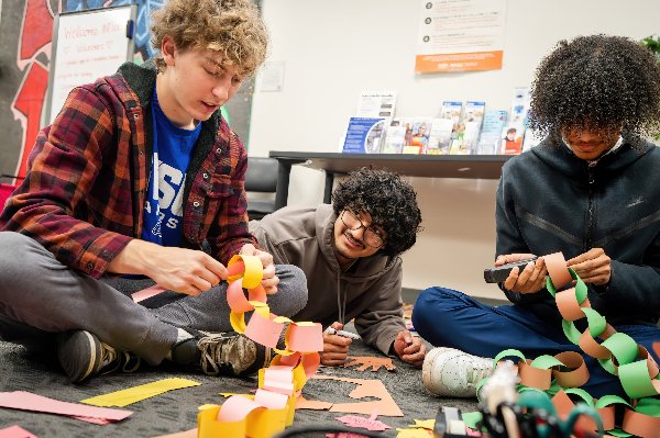 Three young men sit on the floor and assemble a paper chain. 
