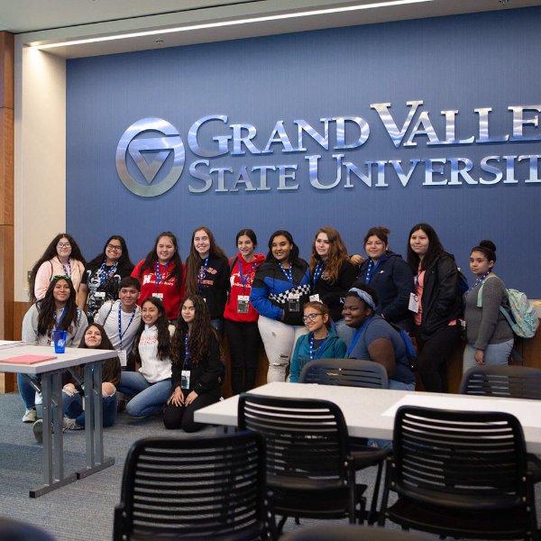 In this 2019 photo, two rows of Latino youth pose for a photo against a blue wall with the GVSU logo and words: Grand Valley State University