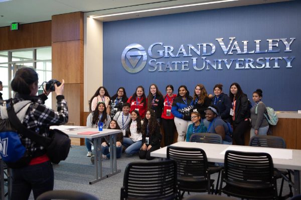 in this 2019 photo, two rows of Latino youth pose for a photo against a blue wall with the GVSU logo and words: Grand Valley State University