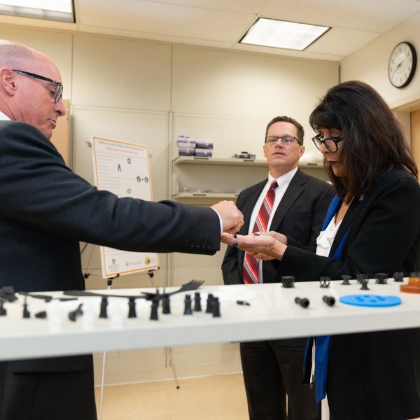 Brent Nowak hands a 3D piece to President Mantella while John Hall looks on. They are in the aMDI labs in the Cook-DeVos Center for Health Sciences.