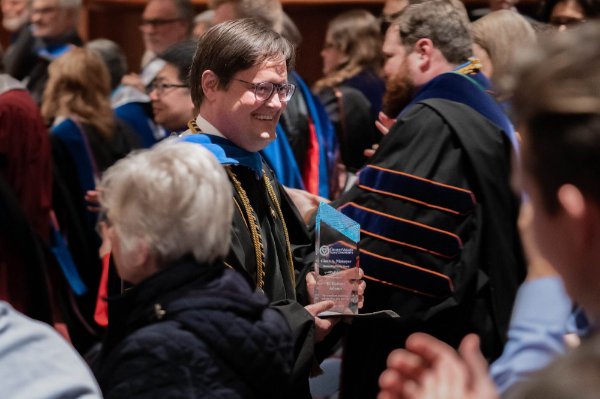 Robert Adams holds an award in a crowd of people, some are wearing academic regalia