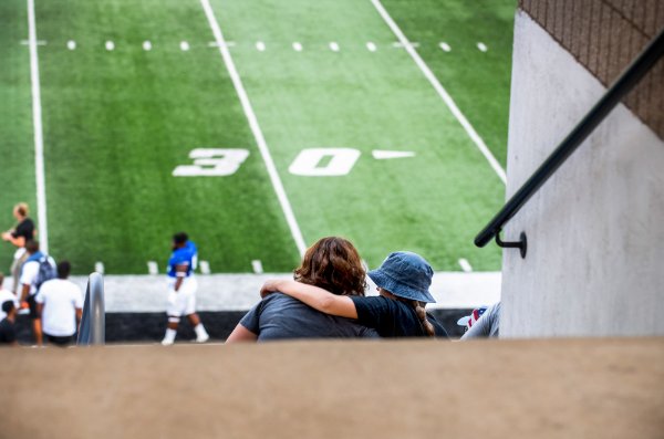 A view from behind shows two people hugging with a football field in the background.