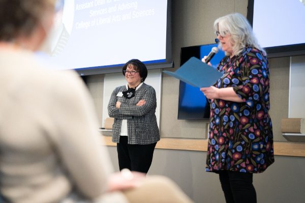 Monica Johnstone, right, holds a microphone and speaks to the audience about Betty Schaner, standing at left. A person is seated in the foreground.
