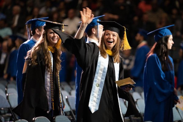 Graduate smiles and waves at someone in the crowd at commencement.