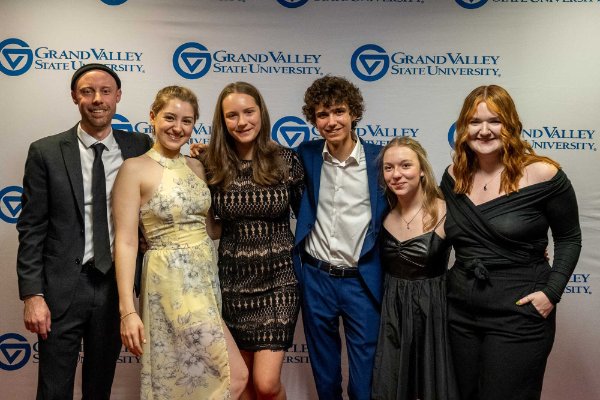 Six people stand side by side, smiling, in a posed shot with a backdrop containing the Grand Valley State University logo.