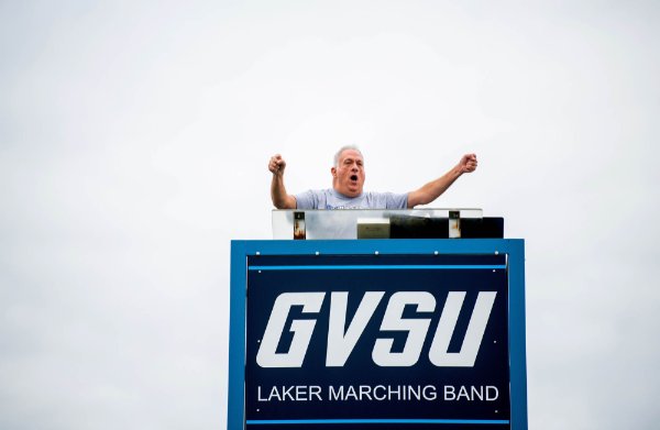 A person gestures while directing a band from a podium that contains a sign with the words, "GVSU Laker Marching Band."