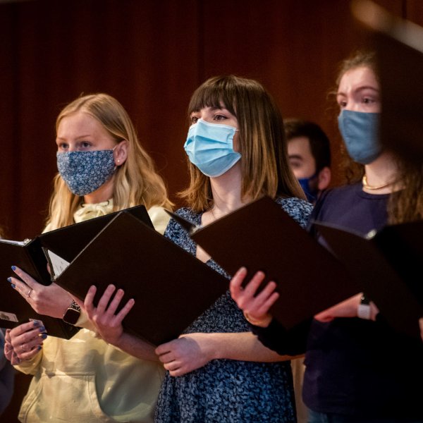 Several people, wearing masks and holding notebooks, sing as part of a choir.