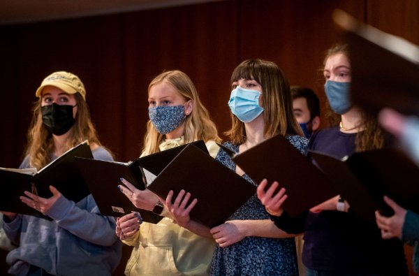 Members of a choir, wearing masks and holding notebooks, sing.