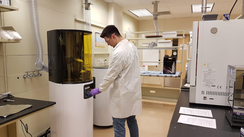 A technician in a lab coat stands in a lab in front of a Carbon, Inc., 3D printer. The printer is a tall cylinder with an amber plastic shield in front of it.