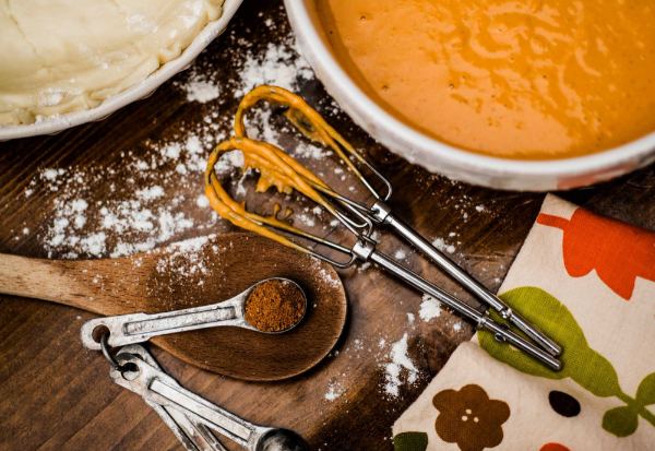 A pie crush, a pumpkin pie, a measuring spoon filled with nutmeg and beaters with pie filling on a counter with flour scattered