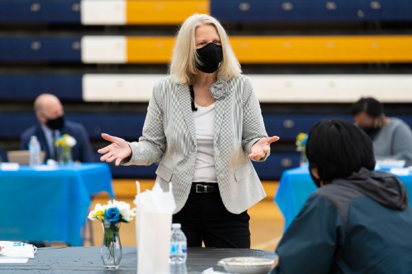 Jean Nagelkerk, with mask on, stands and chats with a student seated at a table