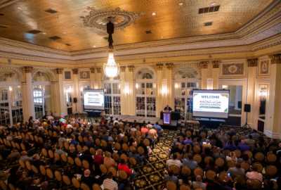 large crowd in ballroom seated with speaker and podium at front, 2 projection screens, large light fixture hanging from ceiling