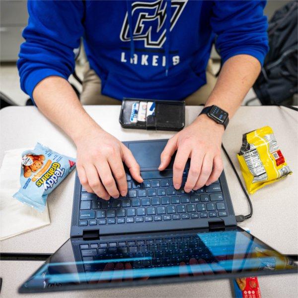 Overhead view of a person wearing a blue Grand Valley Lakers sweatshirt typing on a laptop at a table, with snack bags, a water bottle and a wallet nearby.