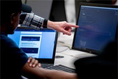 A person points to lines of code on a desktop monitor while another looks on, with a laptop and keyboard on the desk, suggesting a collaborative cybersecurity or programming session.