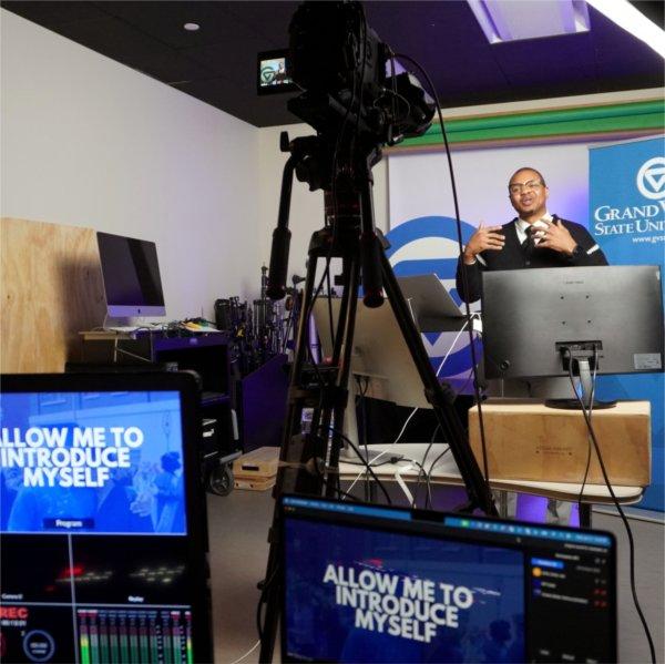 Dar Mayweather in a studio with camera and monitors, he is wearing a dark sweater and white shirt with black tie, with glasses