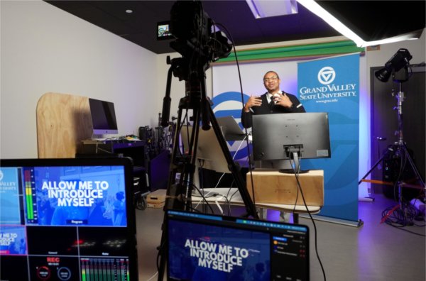 Dar Mayweather in a studio with camera, lights and monitors, he is wearing glasses, black sweater and white shirt