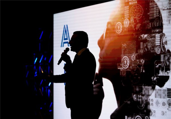 A silhouette of a person using a microphone stands in front of a screen showing the profile of a woman with tech symbols overlaid. 