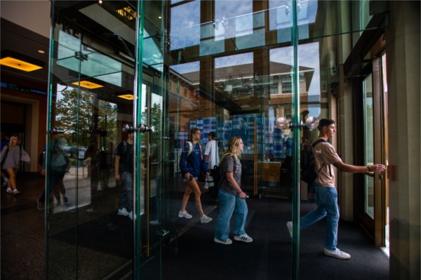Students walk through a glass vestibule on their way out of a campus academic building. 