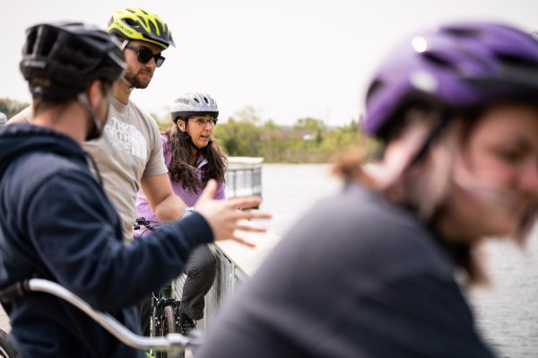four people in bike helmets look at a lake while standing on a boardwalk