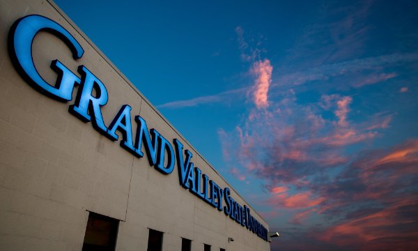 Lit up blue letters spell out "Grand Valley State University" on the side of a tan building against a deep blue sky with pink and purple lit clouds. 