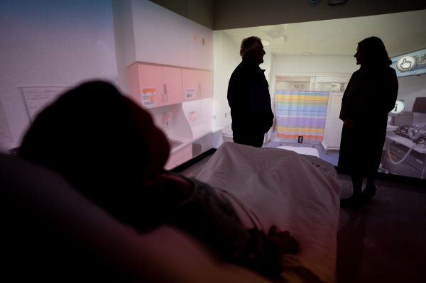 Two people are silhouetted in the background against a projection wall, while a tech-filled manikin used in simulations lays in a hospital bed in the background.