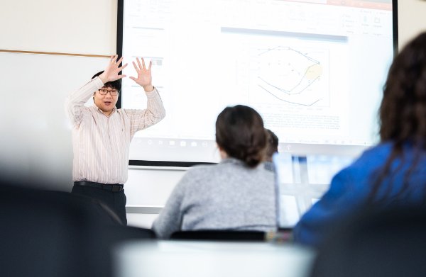 A professor stands at the front of a classroom gesturing with hands above his head while students look on. 