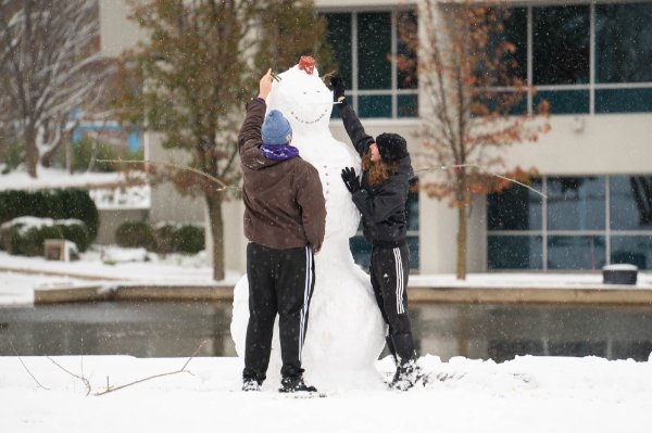 Two people are putting the finishing touches on a snowman with a building and pond behind them. 