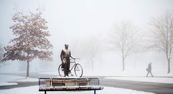 A person walks by a statue of a person with a bike. It is foggy and the ground is covered in a light dusting of snow. 