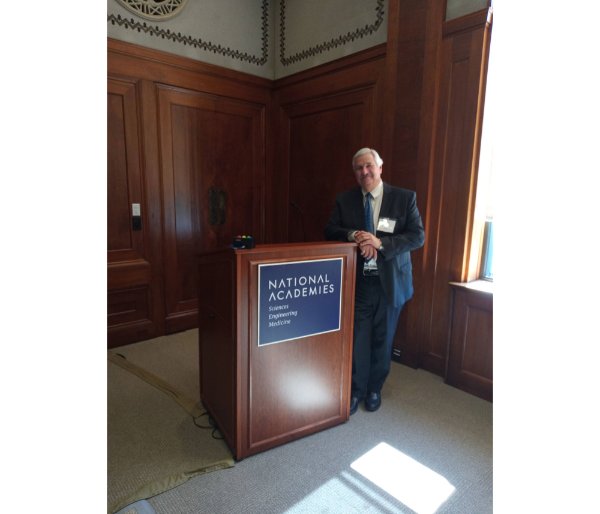 Paul Plotkowski leans against podium with National Academies, Sciences, Engineering, Medicine on front of podium on blue plague 