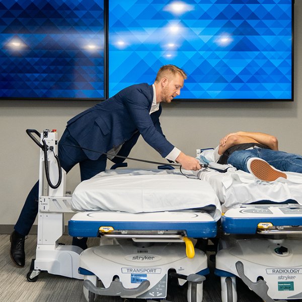 Andrew Heuerman in a blue suit demonstrates the SimPull project by latching straps underneath a mock patient on a bed to be pulled to another bed, as in a transfer.