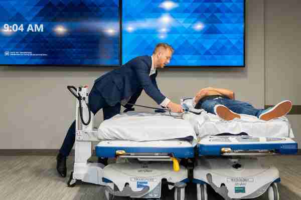 Andrew Heuerman in blue suit latches straps from the SimPull underneath a mock patient to pull the patient to another bed, like a transfer.