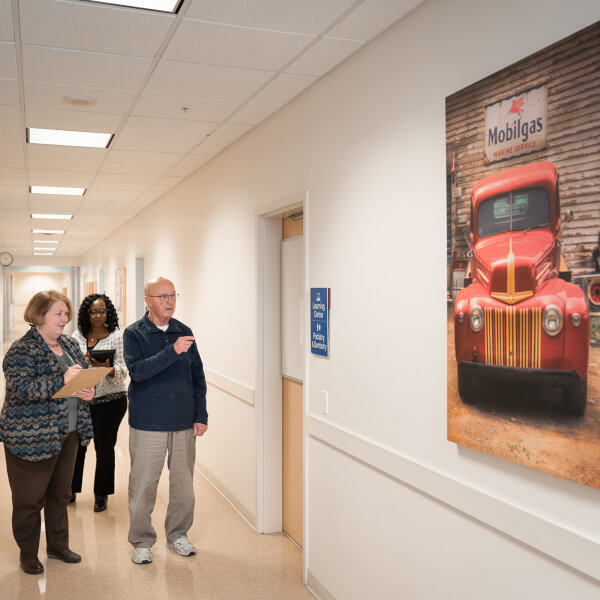 older man and two women looking at large painting of red car and gas station sign