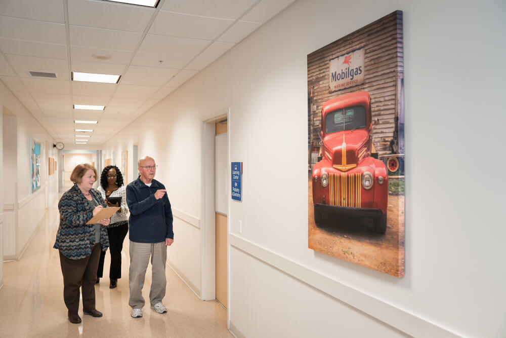 older man and two women looking at large painting of red car and gas station sign