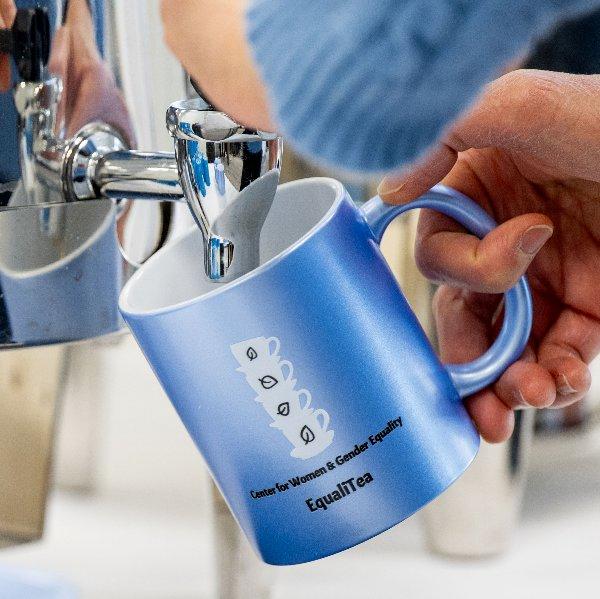 A person pours tea from a coffee urn into a blue mug with text on it that reads �Center for Women and Gender Equity, Equali-Tea�.