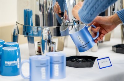 A person pours tea from a coffee urn into a blue mug with text on it that reads &#8220;Center for Women and Gender Equity, Equali-Tea&#8221;. 