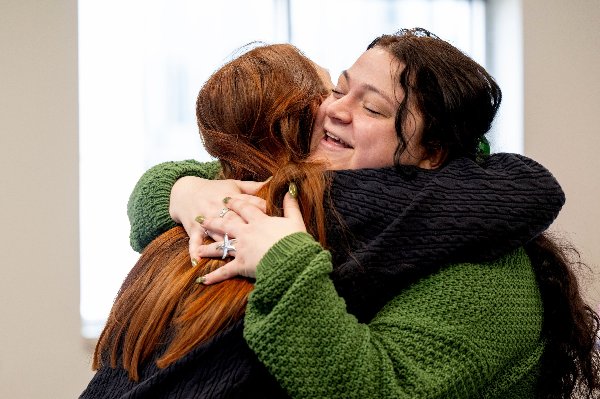 A young woman in a green sweater smiles as she hugs another person.