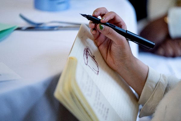 A person sketches a teacup with a purple pen on a blank notebook page. 